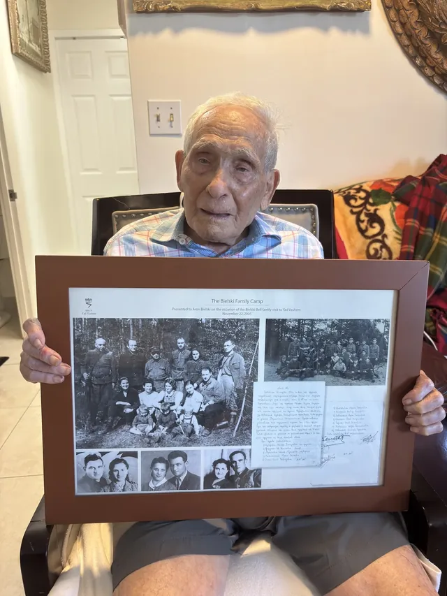 Aron Bielski (Bell) holding a framed display about the Bielski family camp.