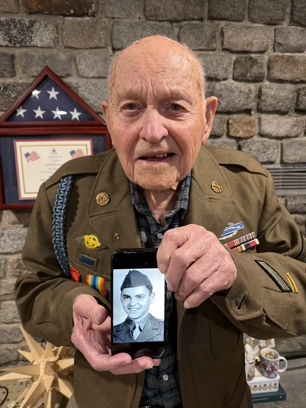 Bud Gahs holding a phone displaying a photograph of himself in Army uniform.
