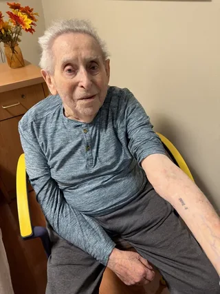 Meir Usherovitz seated and showing the tattoo on his forearm.